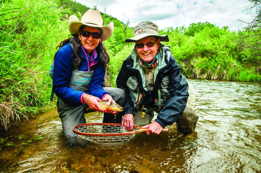 Two women with fly fishing gear kneel in a river